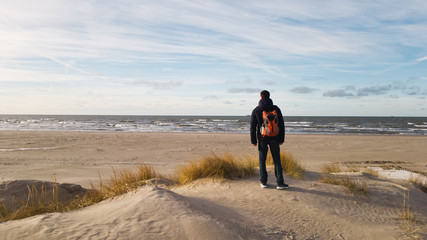 A young man traveler with a backpack stands on the beach and admires the view. Thinking about the future alone. Beautiful light,sand dunes of the cold sea and panoramic views of the endless Baltic sea