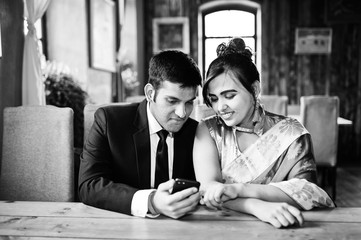 Elegant and fashionable indian friends couple of woman in saree and man in suit posed indoor cafe and looking something on mobile phone.