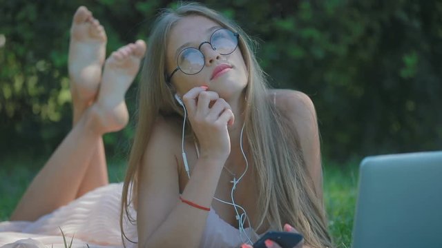 pretty young blond lady in modern glasses and earphones lies and listens to music at laptop on park grass