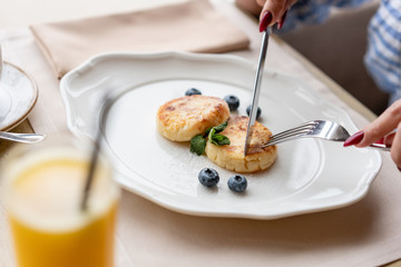 Beautiful young woman cuts pancakes with a knife. Light breakfast with coffee near window in a cafe.