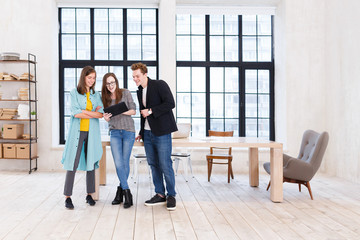 Fototapeta premium Creative teamwork. Positive office workers - two women and man stand near large wooden table and look at the tablet discussing new project on the background of large windows in office