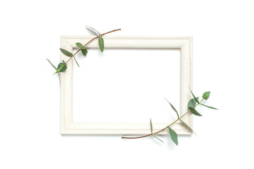 White wooden photo frame and green eucalyptus leaves on white background. Flat lay top view copy space. Stylish minimal composition, artwork mockup, picture frame, home decoration