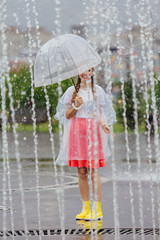 Naklejka premium Young pretty girl with two braids in yellow boots and with transparent umbrella stands near fountain.