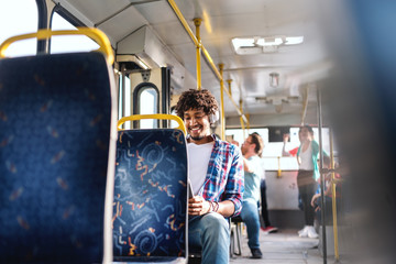 Smiling African American guy sitting in public transportation and using tablet. On ears earphones. In background people sitting and standing.