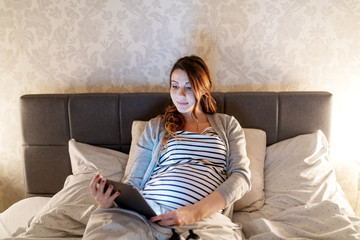 Smiling cute pregnant Caucasian woman with long brown hair lying in the bed and reading about babies on tablet. Evening time.
