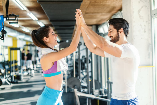 Personal trainer giving high five to sporty Caucasian woman with towel around her neck. Gym interior.
