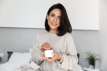Image of adorable woman 30s holding jar with face cream, in modern bright room