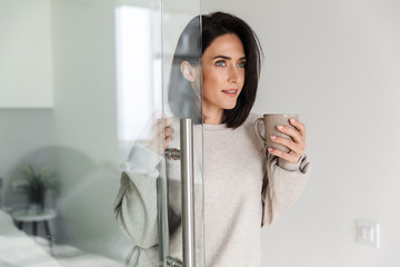 Image of attractive woman 30s holding cup with tea, while standing in modern bright room in the morning