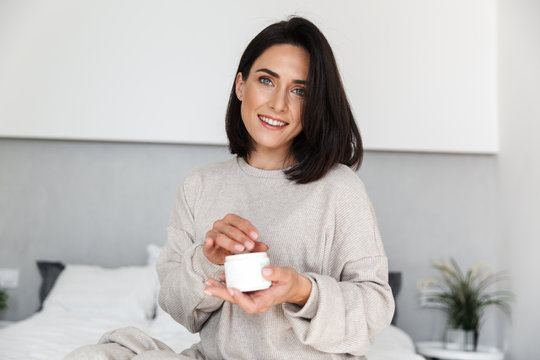 Fototapeta Image of adorable woman 30s holding jar with face cream, in modern bright room