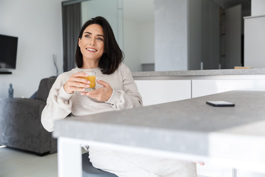 Image of mature woman 30s drinking orange juice, while resting in bright modern room
