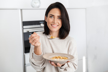Image of brunette woman 30s eating corn flakes on breakfast, while standing in modern kitchen at...