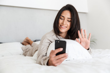 Photo of adult woman 30s using mobile phone, while lying in bed with white linen in bright room