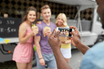 leisure, technology and people concept - young man taking picture of his happy friends eating hamburgers and wok at food truck