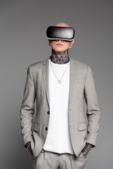 stylish tattooed man standing with hands in pockets and using virtual reality headset isolated on grey