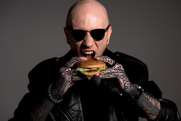 hungry tattooed man in leather jacket and sunglasses eating hamburger isolated on grey