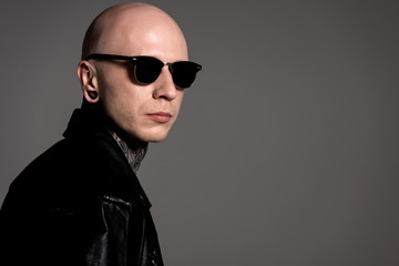 stylish tattooed guy in leather jacket and sunglasses posing isolated on grey