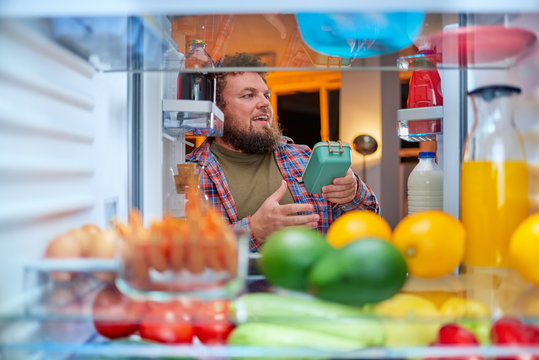  Man looking something to eat at night while standing in front of opened fridge. Unhealthy eating concept. Picture taken from the inside of fridge.