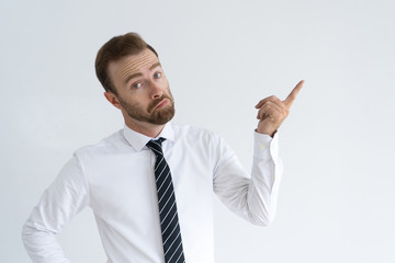Surprised young businessman pointing with forefinger aside while having question. Astonished frowning man in white shirt looking at camera. Advertising concept