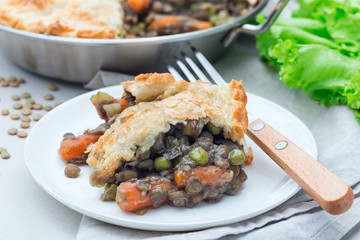Vegetarian pot pie with lentil, mushrooms, potato, carrot and green peas, covered with puff pastry, on a white plate, horizontal