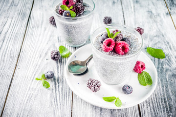 Summer healthy breakfast dessert in glass, overnight vegan chia pudding with non-dairy milk, frozen raspberry, blackberry and blueberry, on light blue and wooden background copy space