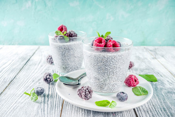 Summer healthy breakfast dessert in glass, overnight vegan chia pudding with non-dairy milk, frozen raspberry, blackberry and blueberry, on light blue and wooden background copy space