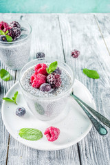 Summer healthy breakfast dessert in glass, overnight vegan chia pudding with non-dairy milk, frozen raspberry, blackberry and blueberry, on light blue and wooden background copy space