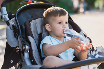 handsome boy sits in a stroller and drinks water from a bottle on a hot summer day