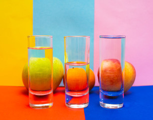 Three glasses of water in the foreground and two apples with mandarin in the background in multicolored tones