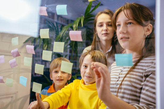 Four different ages pupils with adult female student learning something from colorful stickers glued on glass wall during collaborative process.