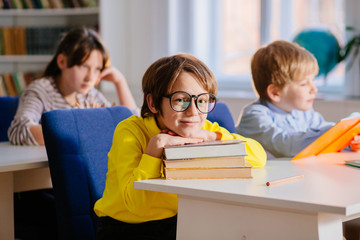 Fototapeta na wymiar Positive school boy in eyeglasses, yellow shirt sitting on desk in classroom with a books. Three school children at desks in elementary school lesson at classroom.