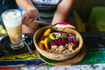 Woman's hands with smoothie bowl made with mango, banana, granola, grated coconut, dragon fruit and mint with latte coffee