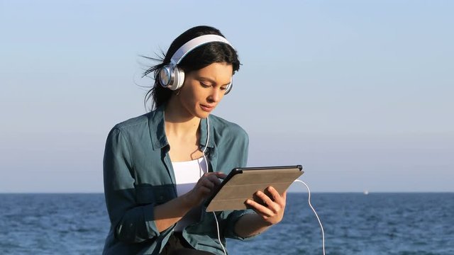 Serious woman browsing and listening tablet content sitting on the beach