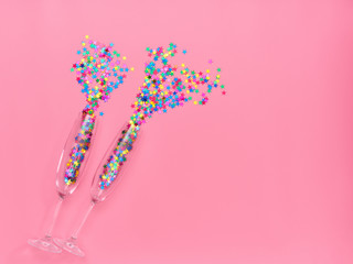 Two champagne glasses with colorful confetti  on pink background. Top view