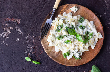 Pasta (farfalle) with creamy sauce with basil and gorgonzola cheese on a wooden plate. Dark brown concrete background. Copy space.