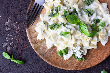 Pasta (farfalle) with creamy sauce with basil and gorgonzola cheese on a wooden plate. Dark brown concrete background. Copy space.