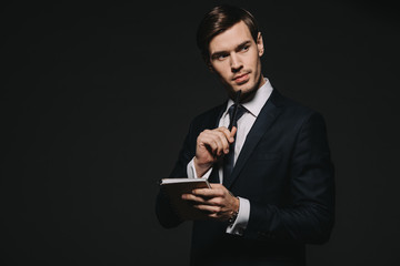 thoughtful businessman holding notebook and pen in hands isolated on black