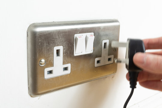 Stainless steel UK plug socket on white wall with hand plugging something in