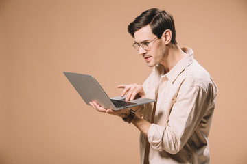 focused man in glasses using laptop isolated on beige