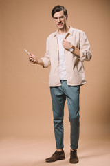 handsome man in glasses holding smartphone and listening music in earphones on beige background