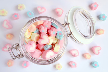 Top View of Pastel Color Heart and Flower Shaped Marshmallow Candies in a Glass Jar with Some Scattered on the Pale Blue Table