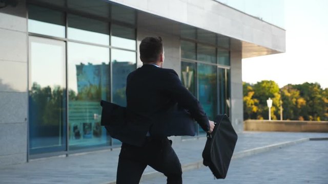 Unrecognizable businessman with briefcase runs down city street. Business man late for meeting. Successful man in suit jogging near modern building. Guy in hurry to appointment. Back view Slow motion