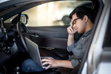 Overworked Asian businessman with glasses feeling stressed and tired while working with laptop computer on driver seat in the car. Migraine symptoms or headache