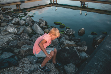 Young blond Caucasian boy pink shirt standing on a dock pointing towards the water during sunset