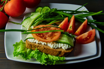sandwich with cheese and sliced tomato cucumbers, lying on a white plate, decorated with green onions, standing on a dark wooden table