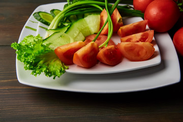 sliced tomatoes cucumbers lying on a white plate decorated with green onions standing on a dark wooden table