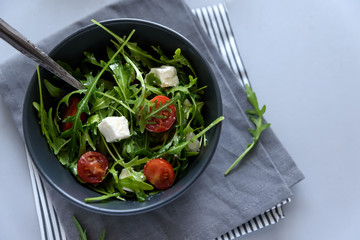 Bowl of arugula salad with tomatoes and cheese on gray wooden background. Vegetarian food concept