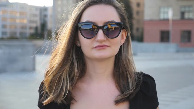 Portrait of young businesswoman in sunglasses walking in city street. Attractive business woman looking at camera. Face of confident girl commuting to work. Blurred background.Close up Slow motion