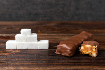 Cubes of sugar in chocolate bar with caramel and peanut on wooden background.