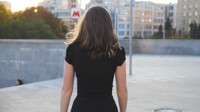 Young unrecognizable girl in black dress walking along urban street. Attractive businesswoman going in city. Girl stepping outdoor. Rear back view Slow motion Close up