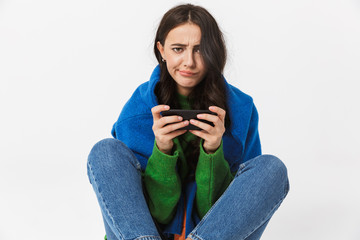 Image of joyous woman 30s in colorful clothes sitting on the floor and using cell phone, isolated over white background